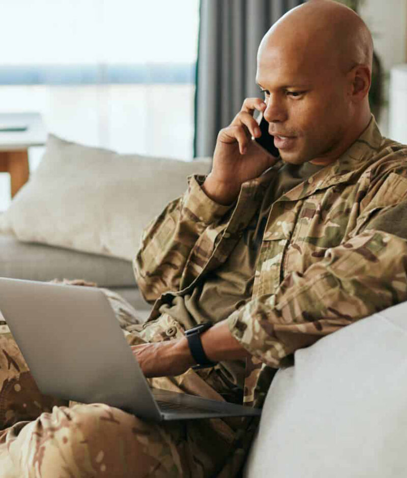 Black military man using laptop while talking on the phone at home. Young African American soldier communicating over cell phone while surfing the net on laptop at home.